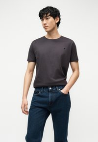 Νεαρός άνδρας που φοράει σκούρο γκρι κοντομάνικο μπλουζάκι και μπλε τζιν, στέκεται με το ένα χέρι στην τσέπη μπροστά σε απλό λευκό φόντο.