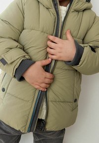 Chaqueta acolchada de color verde oliva con capucha, puños acanalados grises y cierre frontal con cremallera. Presenta costuras a contraste y botones de presión.