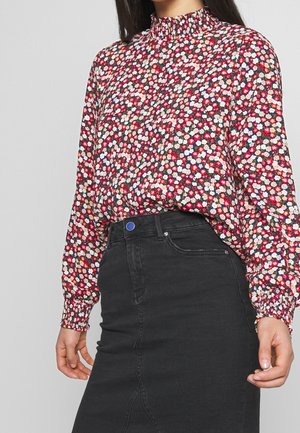 Žena nosící dlouhý rukáv květovanou blůzu s červenými, bílými a růžovými květy, zastrčenou do černé džínové sukně s vysokým pasem.