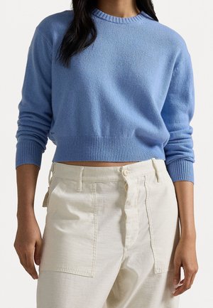 Femme portant un pull court bleu clair et un pantalon taille haute blanc cassé avec de grandes poches avant sur un fond uni.