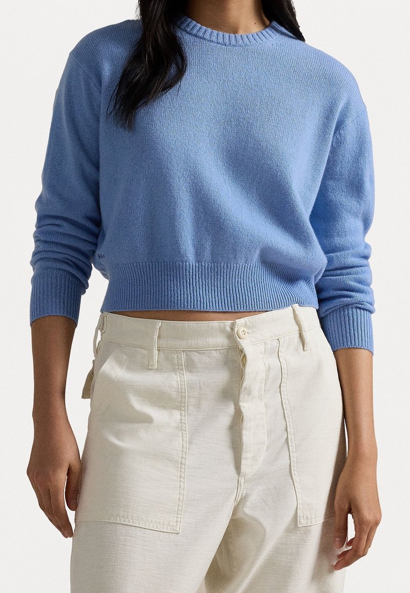 Femme portant un pull court bleu clair et un pantalon taille haute blanc cassé avec de grandes poches avant sur un fond uni.
