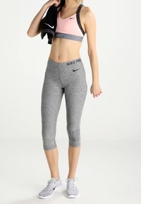 Biustonosz sportowy Nike Pro w kolorze różowym i szarym, zestawiony z szarymi legginsami 3/4 z detalem logo. Model trzyma czarny ręcznik. Szare sneakersy.