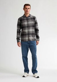 Karierte graue und schwarze Flanelljacke mit zwei Brusttaschen, kombiniert mit blauer Jeans und weißen Turnschuhen, in einer stehenden Pose vor neutralem Hintergrund.