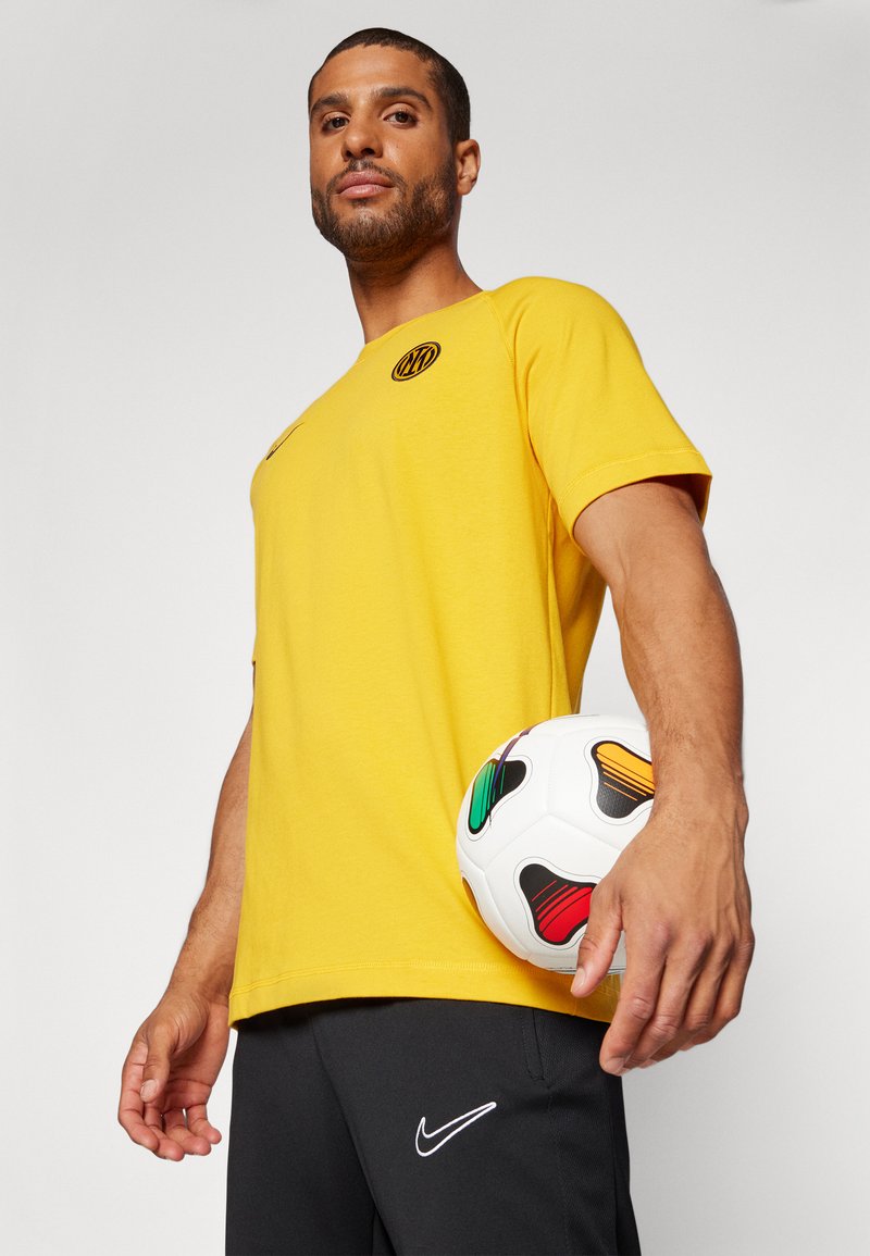 T-shirt sportivo giallo con collo rotondo, maniche corte e logo nero. Il modello tiene in mano un pallone da calcio con motivo, indossando pantaloni corti neri.