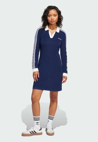 Tamnoplava haljina dugih rukava s bijelim ovratnikom i tri bijele pruge na rukavima. Ima rebrastu teksturu i Adidas logo na prsima.