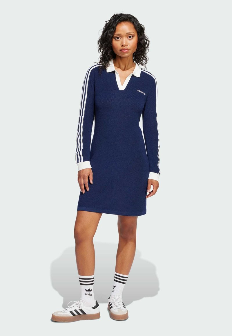 Tamnoplava haljina dugih rukava s bijelim ovratnikom i tri bijele pruge na rukavima. Ima rebrastu teksturu i Adidas logo na prsima.