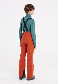 Oranje waterdichte broek met zwarte verstelbare bretels, ontworpen voor buitengebruik, met een losse pasvorm en versterkte onderbenen.