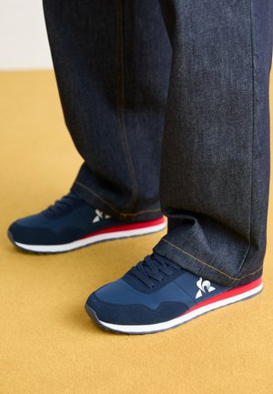 Zapatillas deportivas azul marino con acentos en rojo y blanco, con una parte superior texturizada, cierre de cordones y un logo en el lateral. Combinadas con jeans oscuros.
