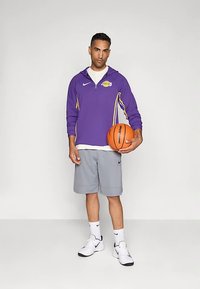 Lila Nike Sweatshirt mit Reißverschluss bis zur Brust und gelben Akzenten, graue Basketball-Shorts, weiße Socken und schwarze Nike-Schuhe, hält einen orangefarbenen Basketball.