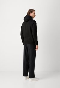 Sweat-shirt noir avec capuche, poignets et ourlet côtelés, associé à un pantalon noir ample et des baskets blanches, vu de dos.