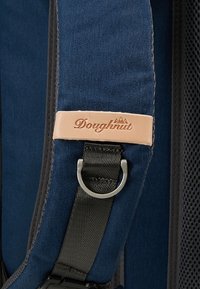 La sangle du sac à dos en toile marine présente une étiquette en cuir avec le logo "Doughnut" embossé, une bague en métal argenté et un dos en mesh noir texturé.