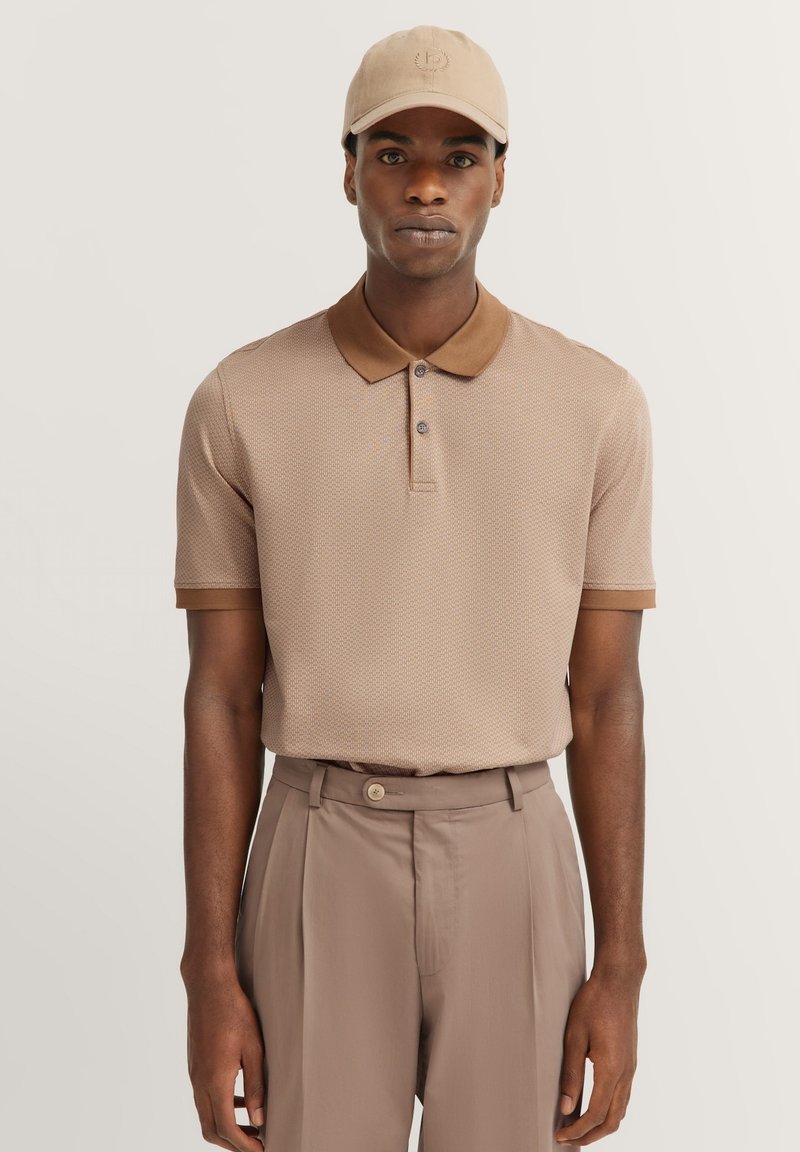Brown textured polo shirt with a classic collar and button placket, paired with light brown trousers featuring a waist button. Beige cap completes the look.