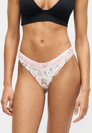 Femme portant un soutien-gorge noir et une culotte à motif floral avec une bordure en dentelle rose, debout, les bras détendus le long du corps.