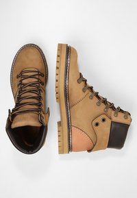 Hnědé kožené kotníkové boty s kovovými doplňky, hnědými akcenty, kulatou špičkou a robustní podrážkou s hlubokým vzorem. Šněrovací design s polstrovaným límcem.