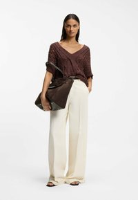 Nainen yllä löysä ruskea neulepaita, leveät kermaan taittavat housut, mustat sandaalit ja hän pitää kädessään isoa ruskeaa mokkanahkasta käsilaukkua, jossa on mustat kahvat.