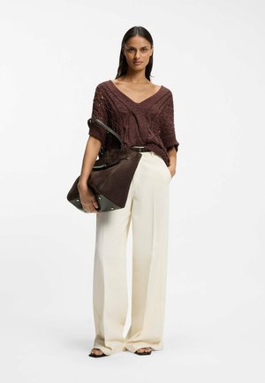 Femme portant un haut en maille marron ample, un pantalon large crème, des sandales noires, et tenant un grand sac à main en daim marron avec des poignées noires.