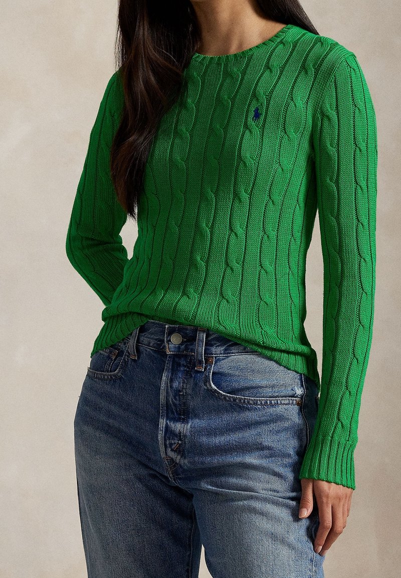 Femme portant un pull vert en maille torsadée avec un petit logo bleu, associé à un jean en denim bleu clair, sur un fond neutre.