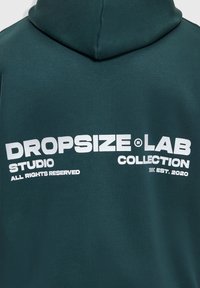 Sweat à capuche vert foncé avec texte blanc indiquant "DROPSIZE LAB STUDIO COLLECTION TOUS DROITS RÉSERVÉS ÉTABLI EN 2020" au dos.