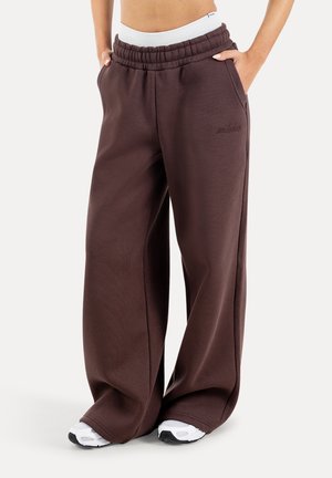 Pantaloni della tuta marrone scuro a gamba larga con vita elastica e tasche laterali, indossati con sneakers bianche e un top corto bianco.