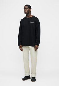 Mann trägt ein schwarzes Langarmshirt, beige Hosen mit Umschlag und schwarze Schuhe, steht vor einem schlichten weißen Hintergrund.