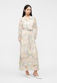 Λουλουδάτο, διάφανο μάξι φόρεμα σε απαλά παστέλ χρώματα με εφαρμοστή μέση, μακριά φουσκωτά μανίκια και στρωματοποιημένο τελείωμα. Συνδυάζεται με λευκά παπούτσια μυτερά στη μύτη.