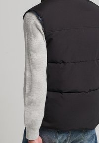 Svart puffy väst med hög krage, bärs över en ljusgrå strukturerad tröja. Vesten har horisontella sömmar och inga synliga knäppningar.