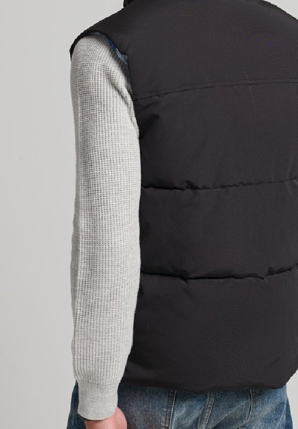 Svart puffy väst med hög krage, bärs över en ljusgrå strukturerad tröja. Vesten har horisontella sömmar och inga synliga knäppningar.