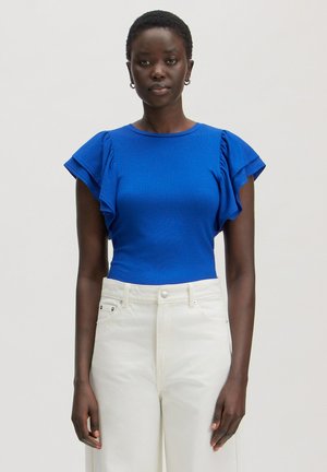 Vrouw met kort haar die een blauw gerimpeld topje met korte mouwen en een witte broek met hoge taille draagt, staand tegen een effen achtergrond.