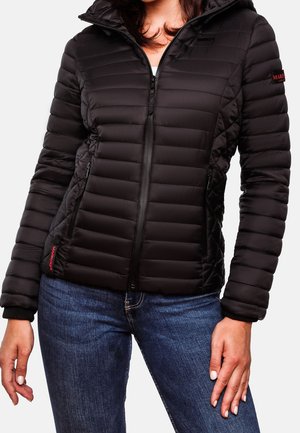Mujer con chaqueta negra acolchada con cremallera y capucha y vaqueros azules, de pie con las manos relajadas a los lados.