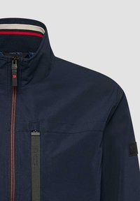 Veste bleu marine avec un col montant, un col intérieur rayé rouge, une fermeture éclair frontale avec accents rouges, et une poche poitrine verticale zippée.