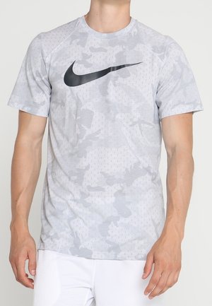 Homme portant un t-shirt Nike camouflage gris clair avec logo swoosh noir et pantalon blanc, debout, les bras détendus le long du corps.