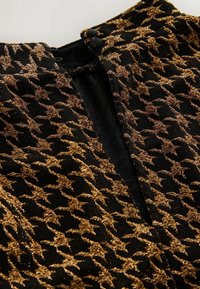 Tissu noir avec un motif pied-de-poule doré scintillant. Présente une fermeture à boutons et une surface texturée. Vue en gros plan mettant en valeur les détails.