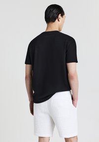 Uomo in piedi con le mani in tasca, che indossa una camicia nera a maniche corte e pantaloni corti bianchi con texture, voltato di spalle su uno sfondo neutro.