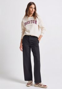 Sweat-shirt crème avec l'inscription "BOSTON 1980" en bordeaux, associé à un pantalon large noir et des baskets marron ornées de touches bleues et rouges.
