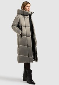 Langer, gesteppter Mantel in Hellgrau mit hohem Kragen, gestepptem Design, seitlichen Taschen und einem Frontreißverschluss. Kombiniert mit schwarzen Stiefeln.