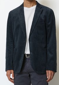 Blazer in corduroy blu navy con sottili strisce verticali, due tasche anteriori, una tasca sul petto e rever a punta, indossato sopra una camicia bianca.