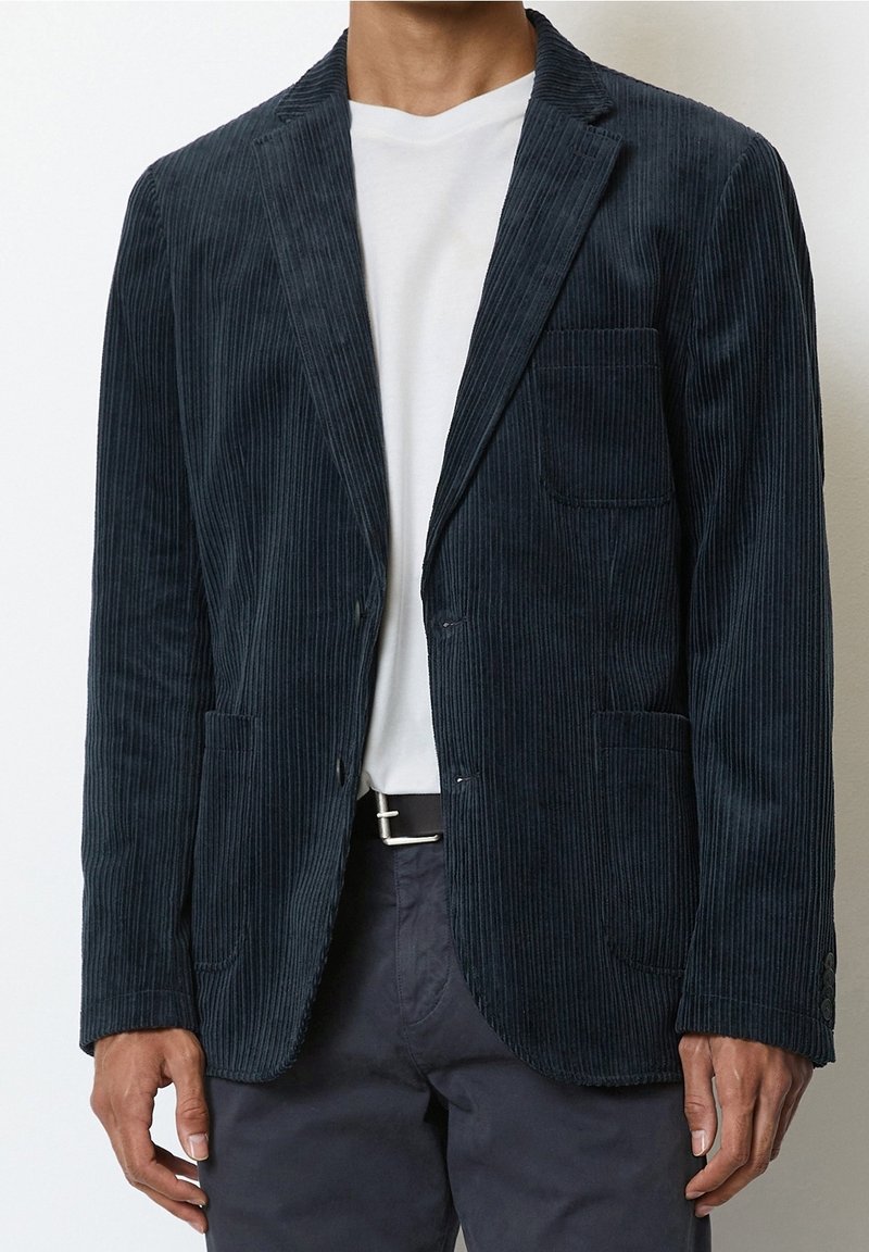 Blazer in corduroy blu navy con sottili strisce verticali, due tasche anteriori, una tasca sul petto e rever a punta, indossato sopra una camicia bianca.