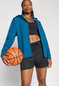 Ženska drži košarkarsko žogo, oblečena v modro jopa s kapuco z zadrgo, črnim športnim nedrčkom in črnimi športnimi kratkimi hlačami, stoji pred enobarvnim ozadjem.