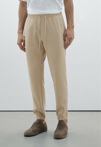 Beige sweatpants met een elastische tailleband, trekkoordsluiting, opgerolde enkels en zijzakken, gecombineerd met bruine suède schoenen.