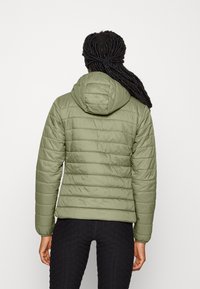 Personne avec de longues dreadlocks portant une veste matelassée à capuche verte clair et un pantalon noir texturé, se tenant de dos face à l'appareil photo.