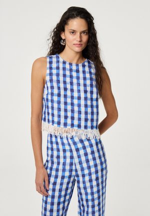 Frau mit langen lockigen Haaren, die ein blau-weiß kariertes ärmelloses Oberteil mit Fransen und passende Hose trägt, steht vor einem einfarbigen Hintergrund.