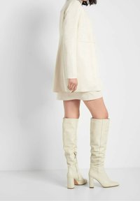 Bottes hauteur genou en ivoire avec une texture en cuir lisse, talons carrés et détail de fermeture éclair latérale. Design simple et épuré avec des accents de couture minimaux.