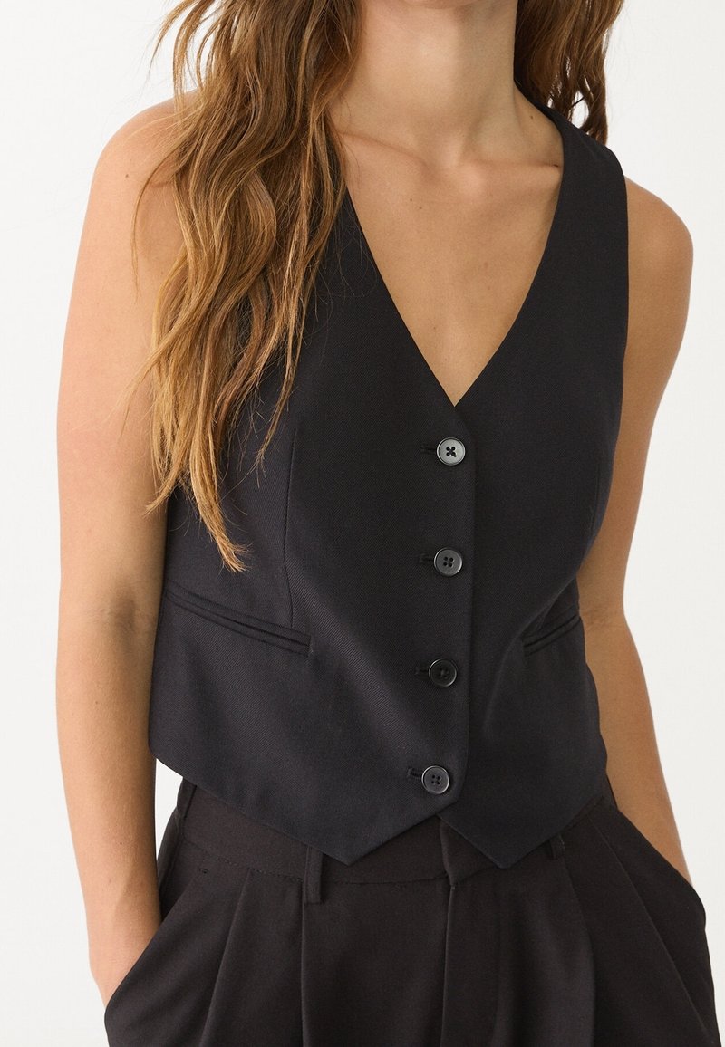 Femme portant un gilet noir sans manches boutonné avec un col en V et un pantalon noir taille haute, les mains dans les poches, sur un fond uni.