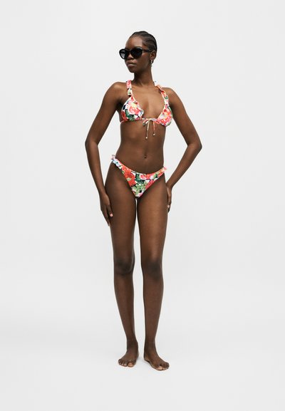 Γυναίκα που φοράει φλοράλ μπικίνι με βολάν στις άκρες και μαύρα γυαλιά ηλίου, στέκεται ξυπόλητη μπροστά σε απλό ανοιχτό φόντο.