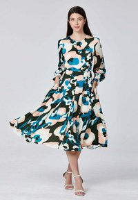 Femme portant une robe fleurie à manches longues, de longueur genou, avec des motifs abstraits bleus, roses, noirs et blancs, assortie à des sandales à talons hauts blanches.