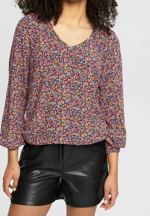 Vrouw die een meerkleurige, langärmelig bloemenblouse met V-hals en zwarte leren shorts draagt, tegen een effen achtergrond.