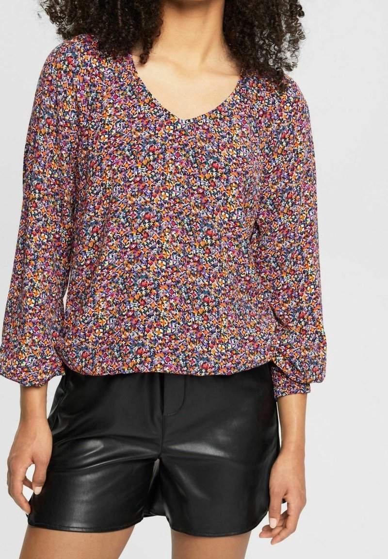 Femme portant un chemisier à manches longues multicolore à motifs floraux avec un col en V et un short en cuir noir, sur un fond uni.