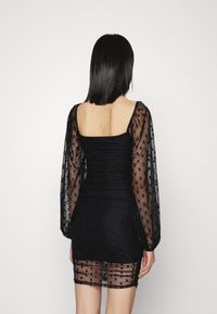 Musta mekko, jossa on sopiva siluetti, neliömäinen pääntie, läpinäkyvät pilkulliset pitkät hihat ja kerrostettu verkkotyyli helman reunassa.