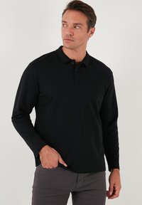 Camisa polo negra de manga larga con cuello, que presenta una tapeta con tres botones. Textura de tela suave, combinada con pantalones grises.