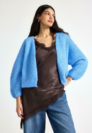 Kvinde iført en lys blå, blød cardigan over en mørkebrun satinbluse med blonder og blå jeans, stående med den ene hånd på hoften.